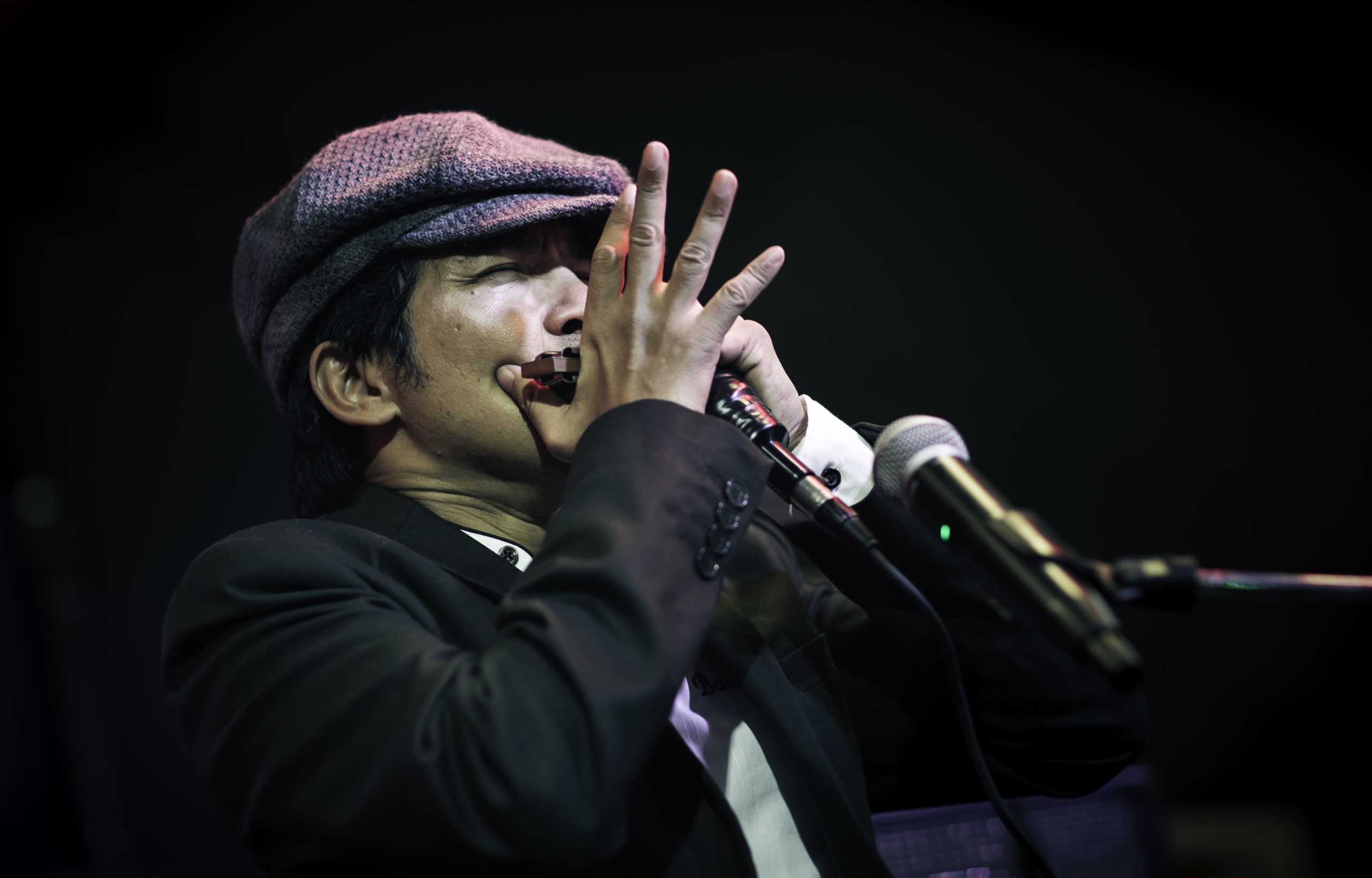 Bonny B der eine Mundharmonika spielt er trägt einen grauen Hut und ein schwarzes Jackett