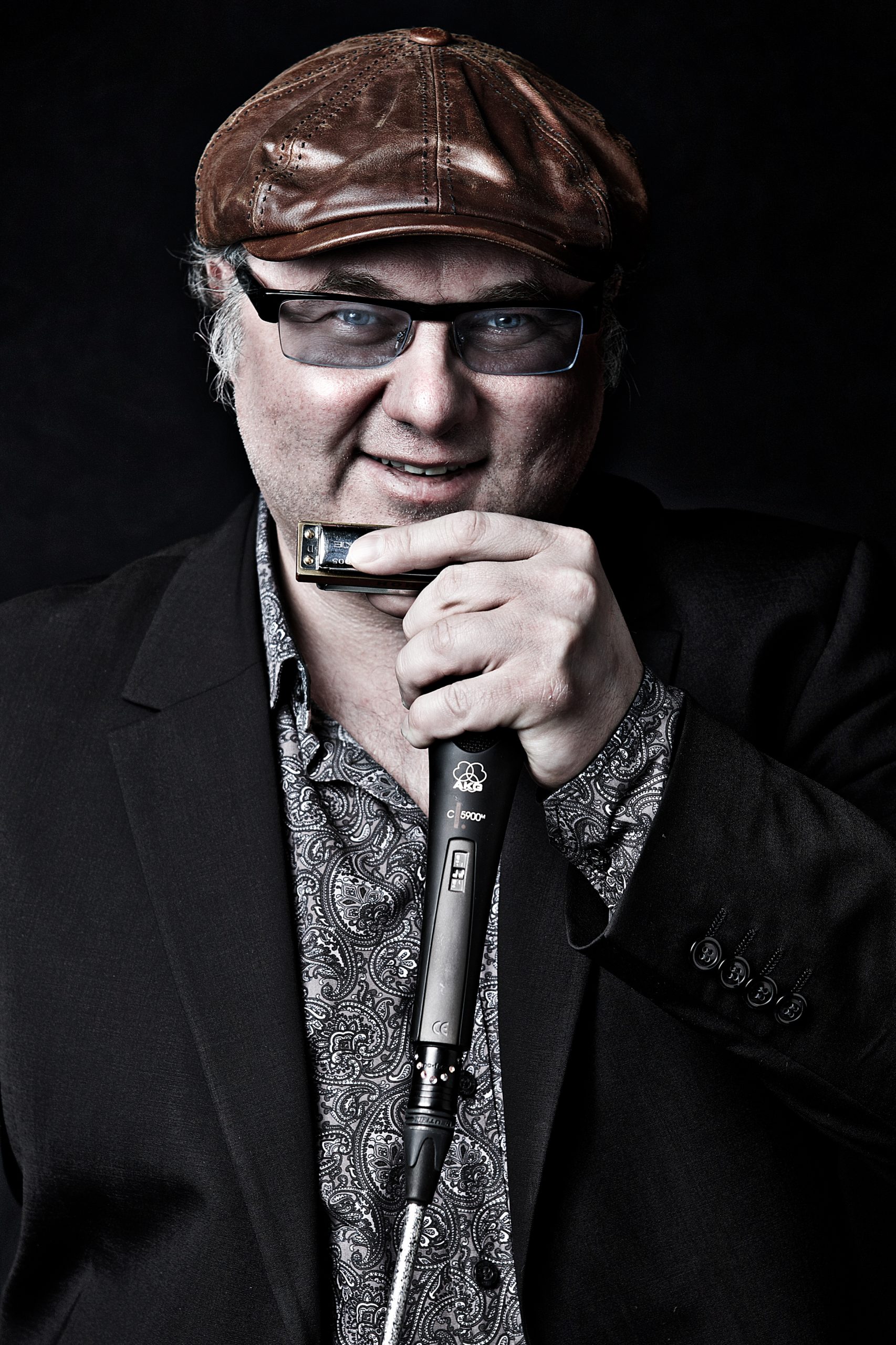 Portrait von Chris Kramer der eine braune Lederkappe trägt und mit der linken Hand ein Mikrofon und eine Mundharmonika hoch hält