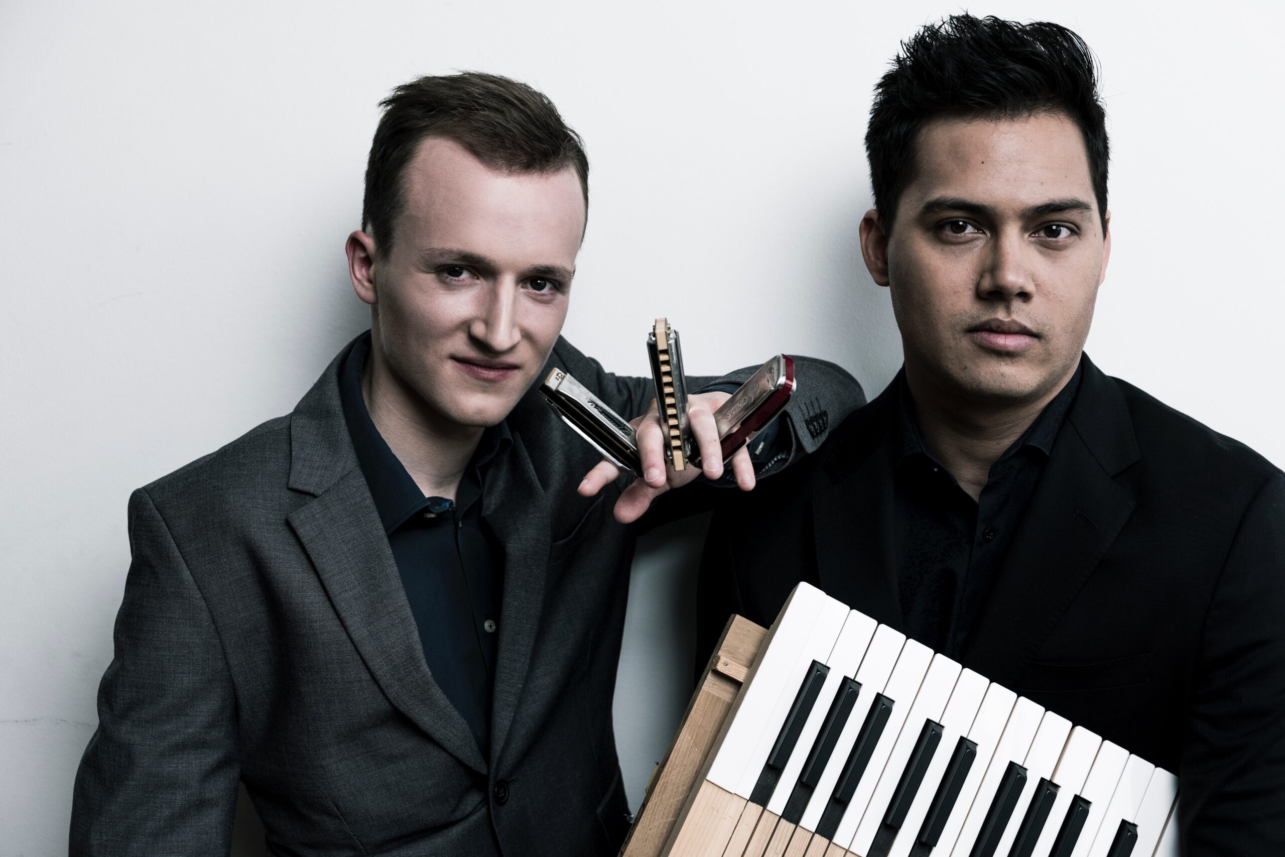 Konstantin Reinfeld und Hanno Busch nebeneinander vor einem weißen Hintergrund. Konstantin Reinfeld hält in der linken Hand drei Mundharmonikas, Hanno Busch ein Akkordeon