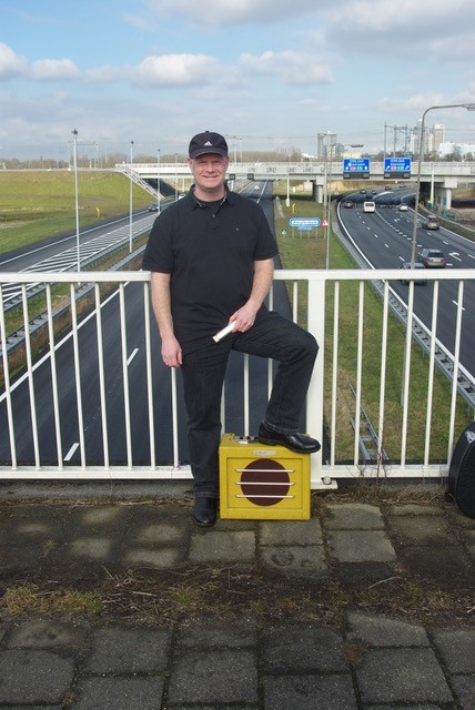 Musiker George Reijnders, fotorafiert auf einer Autobahnbrücke. Er steht mit dem linken Bein auf einem gelben Verstärker, trägt schwarze Jeans und T-Shirt und ein schwarzes Cap