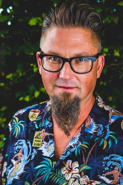 Portrait von Dozent Mattias Bogefors, er träht ein buntes Hemd und eine Brille im Hintergrund sind grüne Blätter